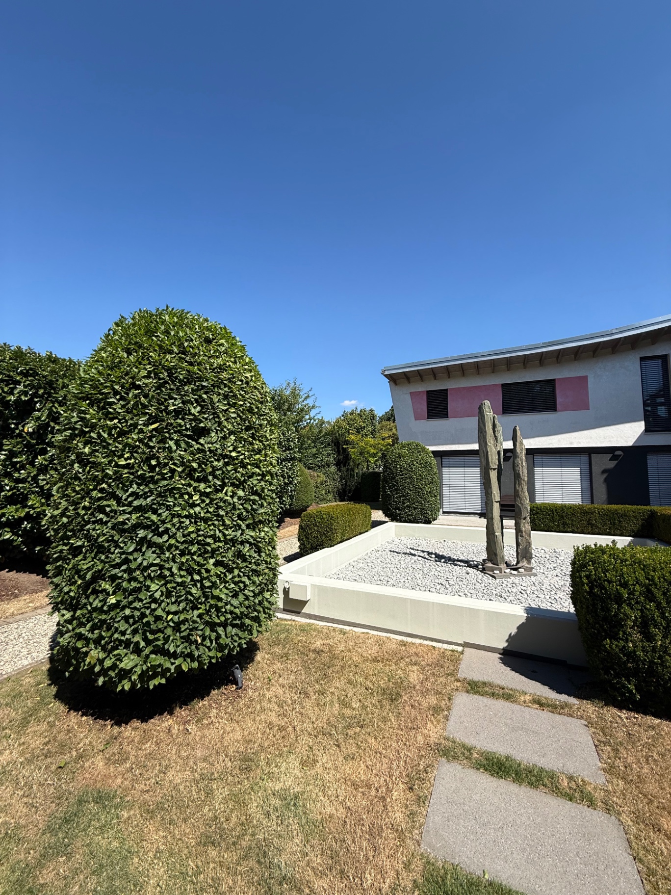 Formgeschnittene Hainbuchen und Buchskugeln neben einem Kiesbeet mit Steinskulptur vor einem modernen Wohnhaus